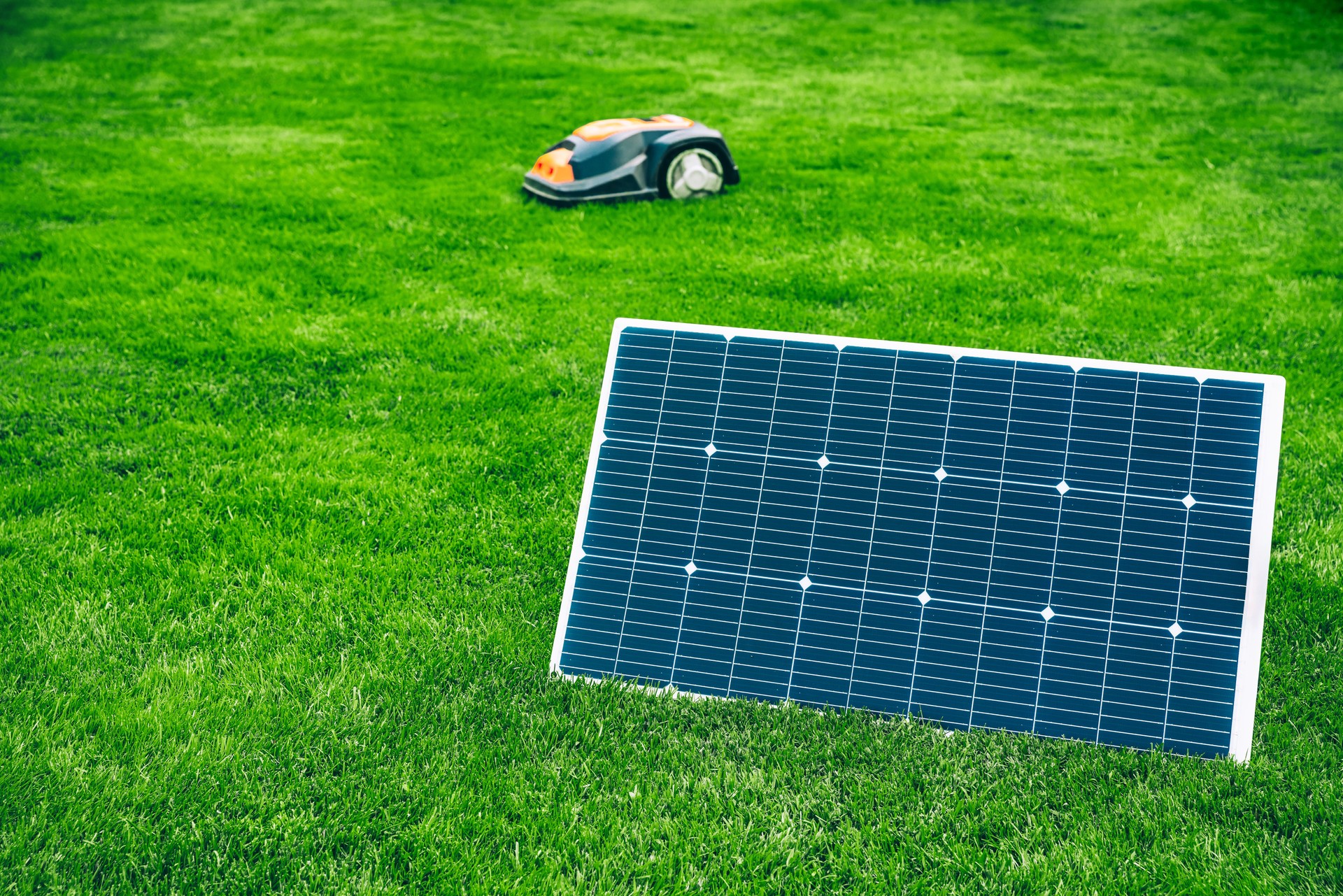 Solar panel generating renewable energy on green grass on a sunny summer day and robotic lawn mower cutting grass in the background.