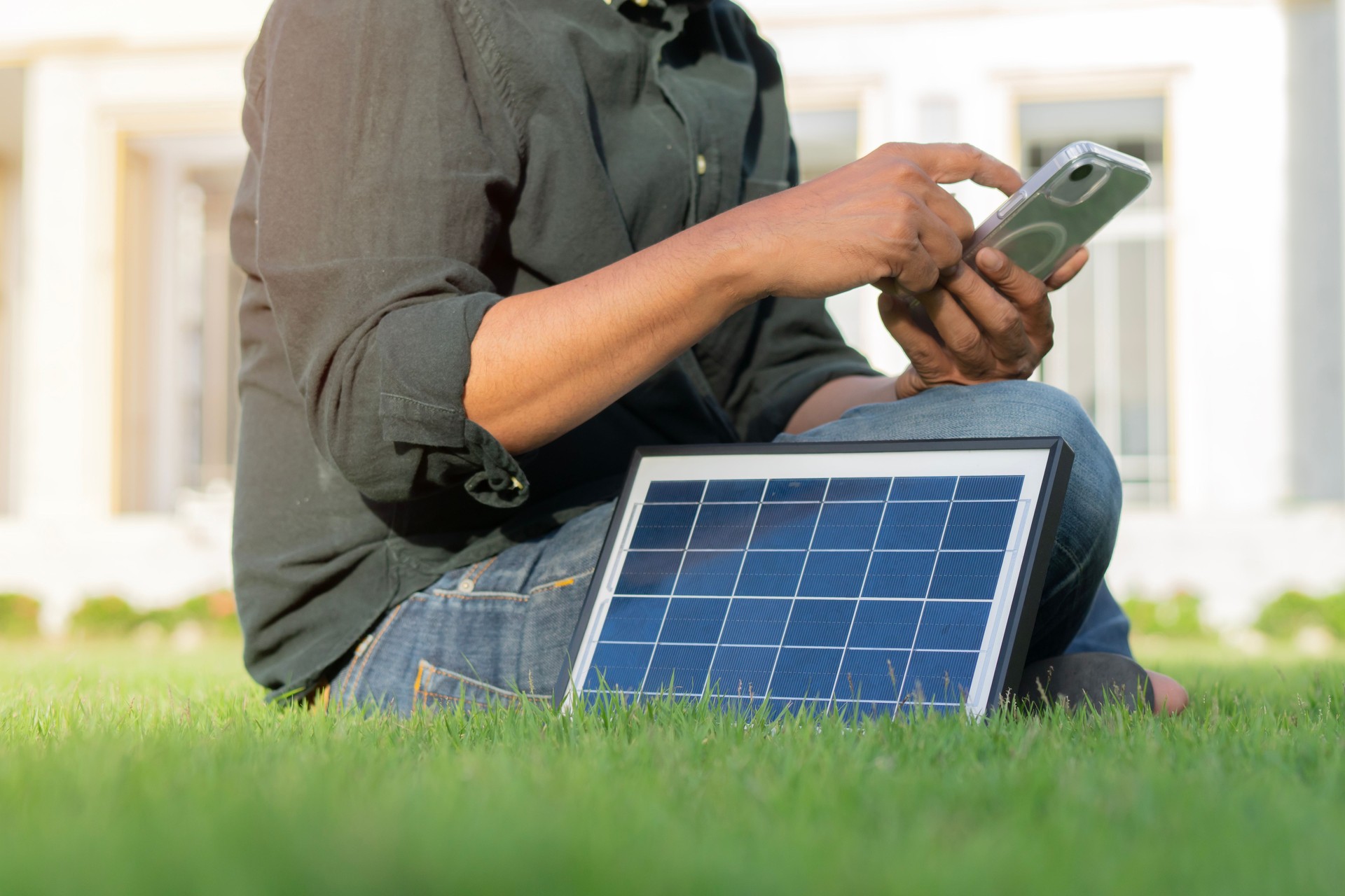 Side view shot of man wearing green casual shirt and sitting on lawn using his smart phone next to mini solar energy panels.